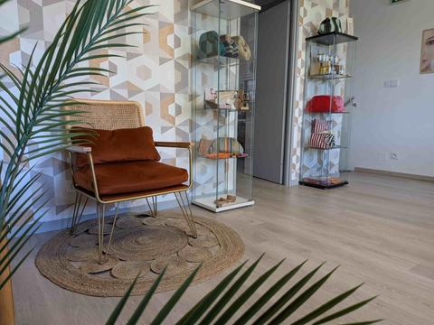 Fauteuil en velours terracotta et cannage devant le papier peint à hexagones or et beige, palmes vertes au premier plan et vitrines en verre garnies d'accessoires