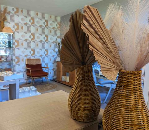 Deux vases en osier tressé garnis de palmes séchées et de pampa sur le comptoir d'accueil, le coin salon et son fauteuil terracotta en arrière-plan