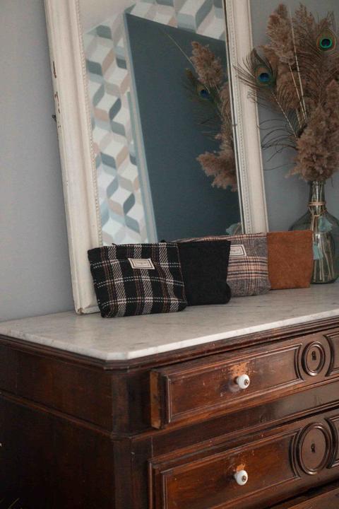 Commode ancienne en bois sombre à plateau de marbre, trousses en tissu à carreaux alignées devant un miroir blanc, plumes de paon et pampa dans un vase