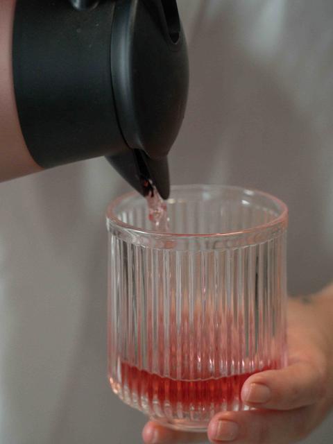 Une main tient un verre strié dans lequel on verse une infusion rouge depuis une verseuse noire