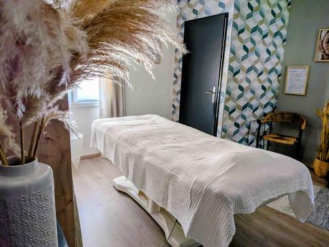 Cabine de soin : table recouverte d'un drap en gaze blanche, papier peint à cubes sauge et gris, fauteuil en rotin et pampa dans un vase au premier plan