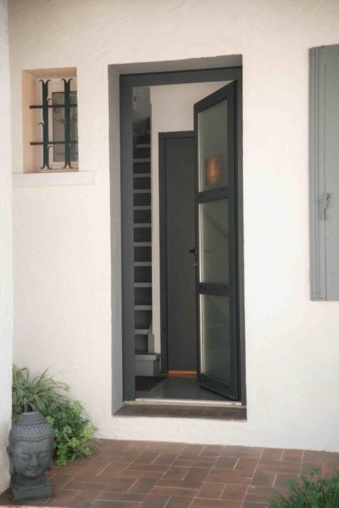 Porte d'entrée vitrée gris anthracite ouverte sur l'escalier qui mène à l'institut, mur blanc crépi, statue de bouddha et plante au pied du perron en tomettes