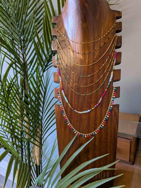 Colliers fins dorés à perles multicolores présentés sur un buste en bois sculpté, feuilles de palmier au premier plan