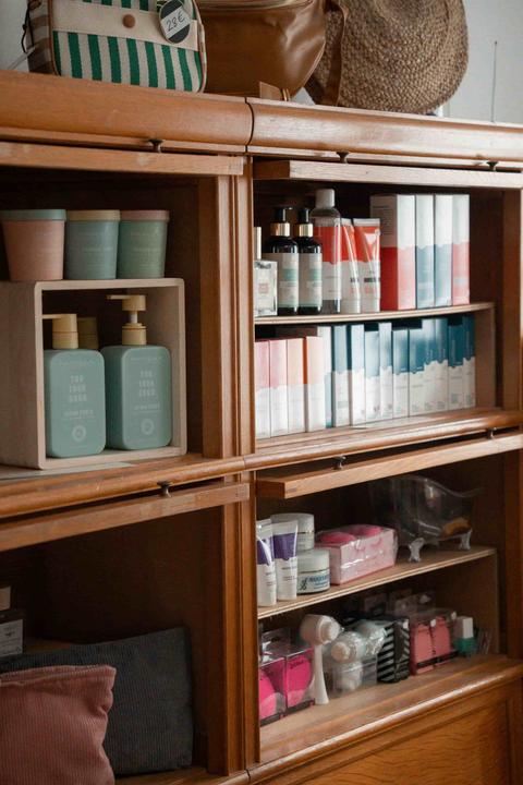 Bibliothèque vitrée en bois blond de l'espace de vente : pots pastel, flacons-pompes vert d'eau Parisax, coffrets de soins blancs, rouges et bleus, sacs et panier de paille posés sur le dessus
