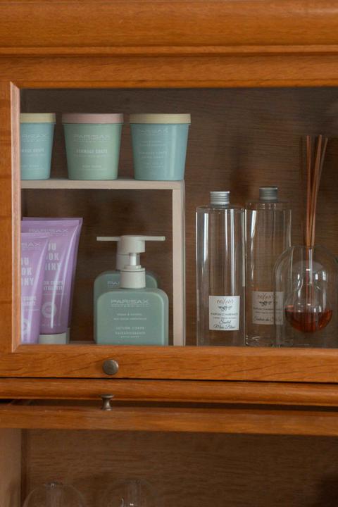 Vitrine en bois : pots vert d'eau et tubes violets de soins pour le corps Parisax, flacons de parfum d'ambiance et diffuseur à bâtonnets