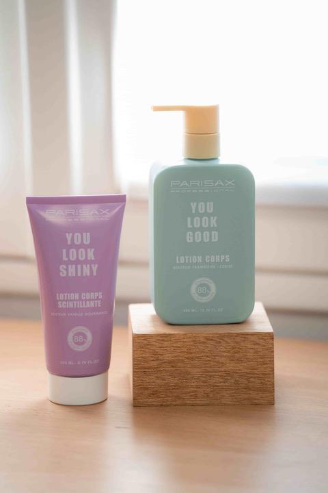 Deux lotions pour le corps Parisax sur une table en bois : un tube violet et un flacon-pompe vert d'eau posé sur un cube de bois