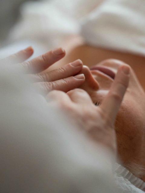 Mains de l'esthéticienne massant le visage d'une cliente allongée, vues à travers un linge blanc flou au premier plan