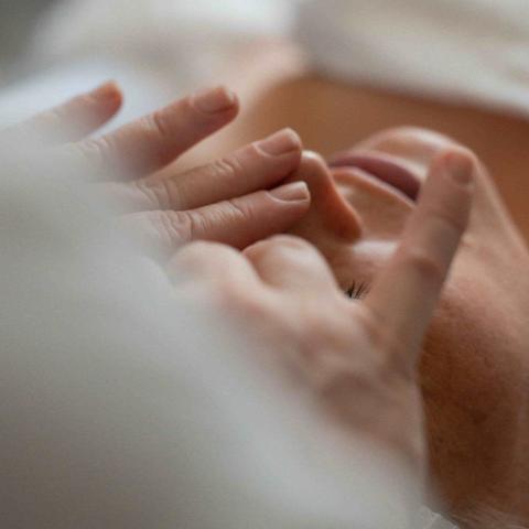 Mains de l'esthéticienne massant le visage d'une cliente allongée, vues à travers un linge blanc flou au premier plan