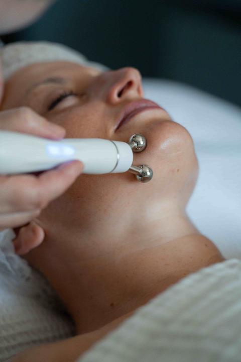 Soin du visage : un appareil à deux billes métalliques glisse le long de la mâchoire d'une cliente allongée, les yeux fermés, sous une serviette gaufrée blanche